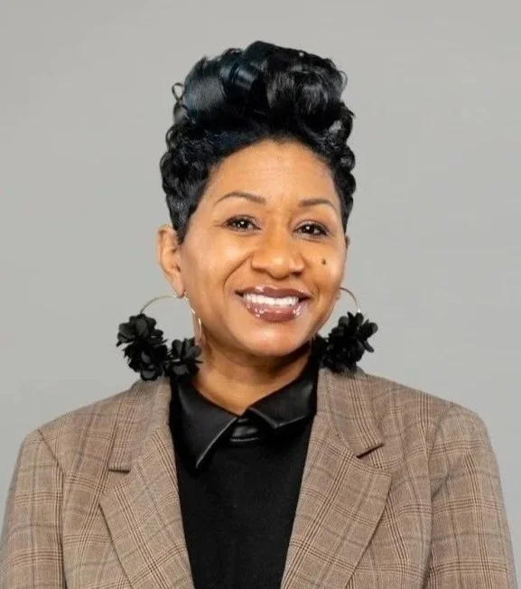 A woman with short, curly black hair styled up, wearing hoop earrings with black flower details, a black shirt, and a brown plaid blazer, smiling against a plain gray background.