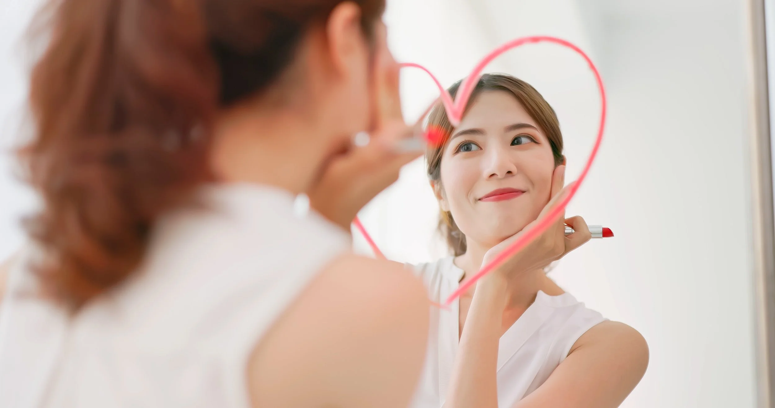 A woman with brown hair and a white top is drawing a red heart on a mirror with a red marker. She is looking at her reflection in the mirror, smiling softly.