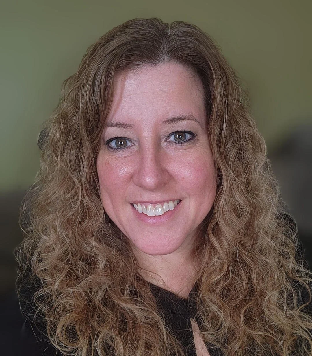 A woman with curly, light brown hair and blue eyes smiling at the camera, wearing a black top, with a blurred green background.