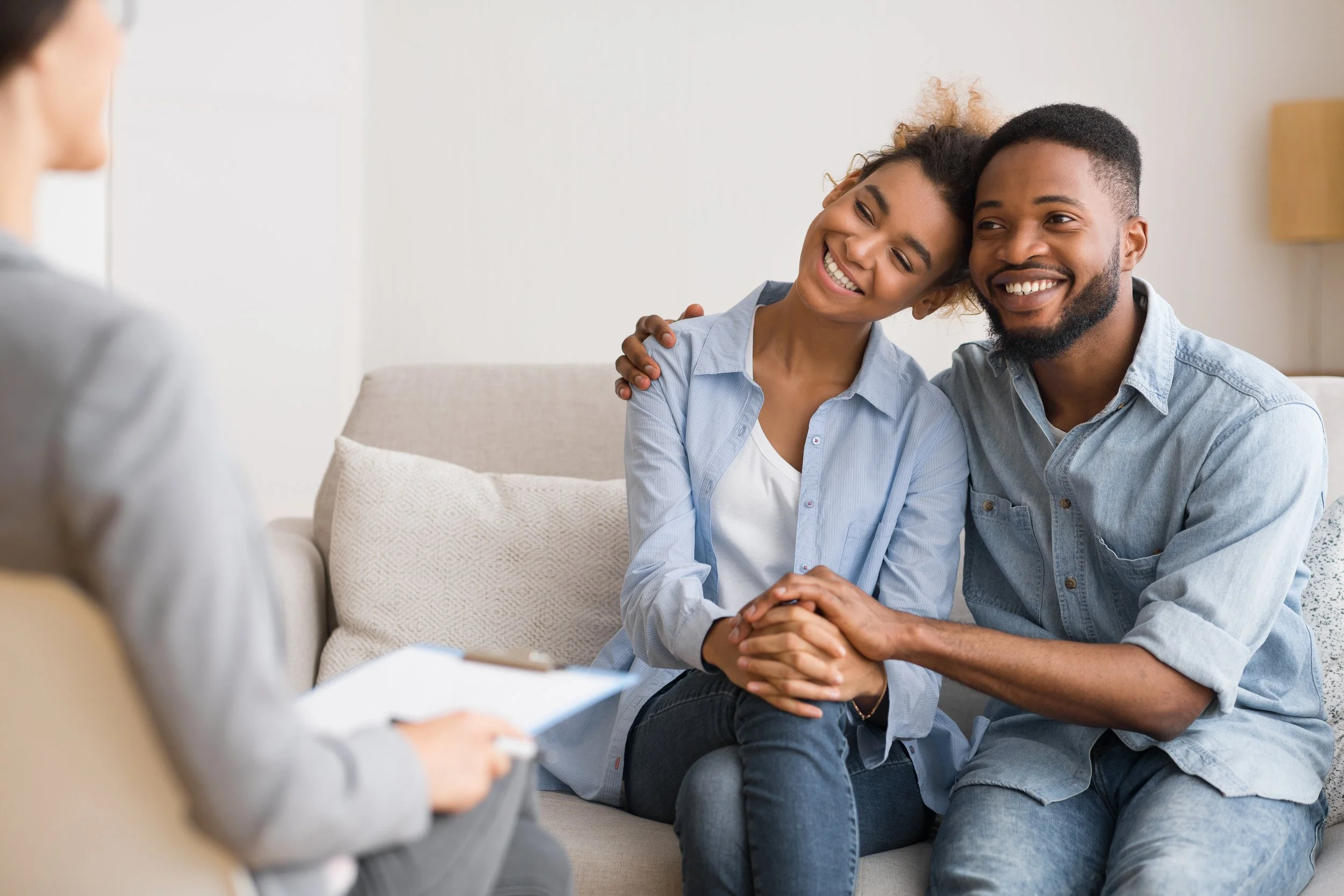 Couple smiling and holding hands during therapy session with therapist taking notes.