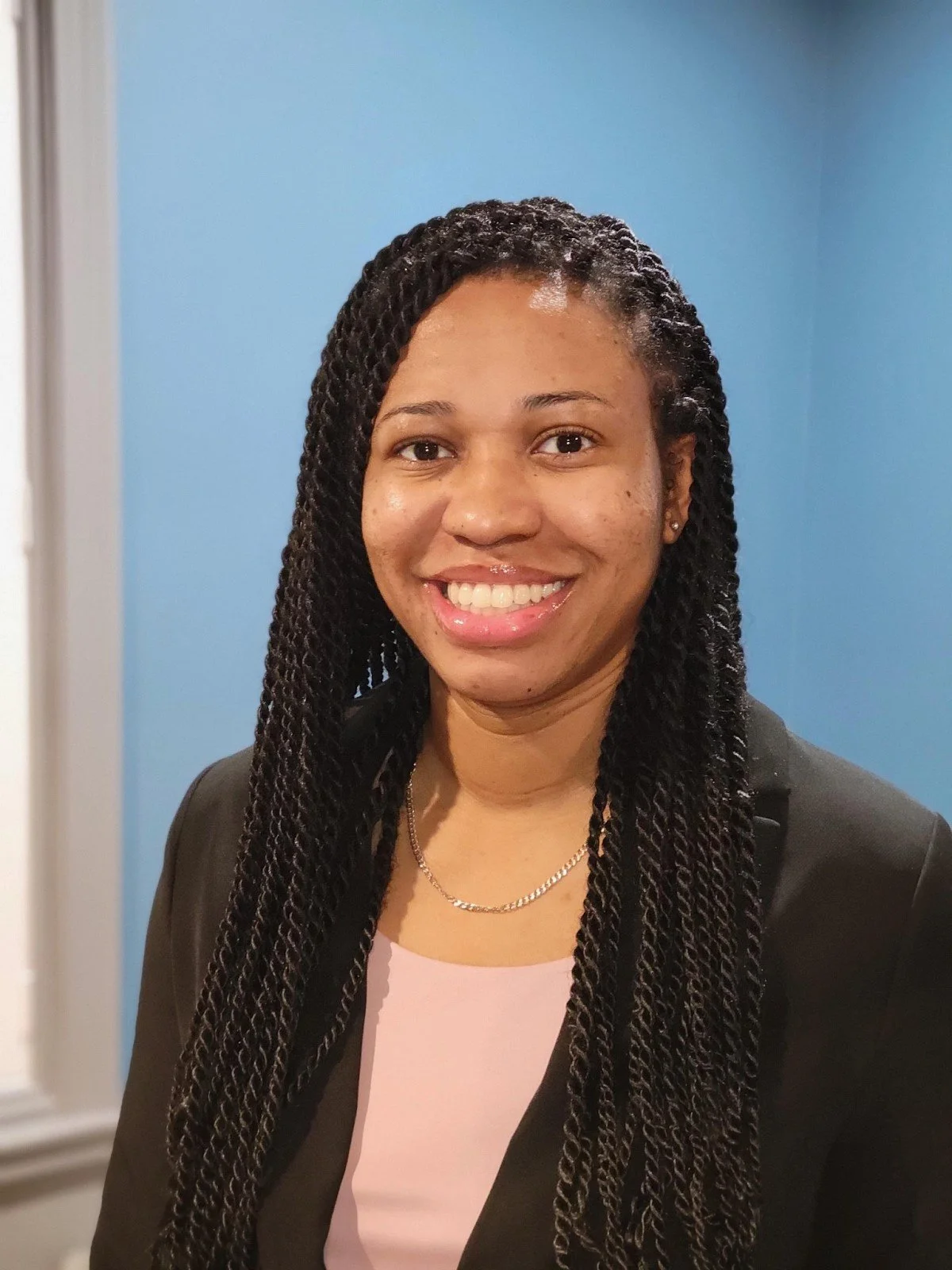 A young woman with braided hair, smiling, wearing a black blazer and a gold necklace, standing in front of a blue wall.