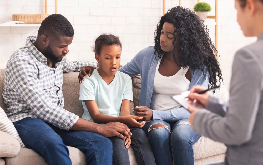 A woman and a man sitting on a couch with a young girl, during a medical appointment with a professional taking notes.