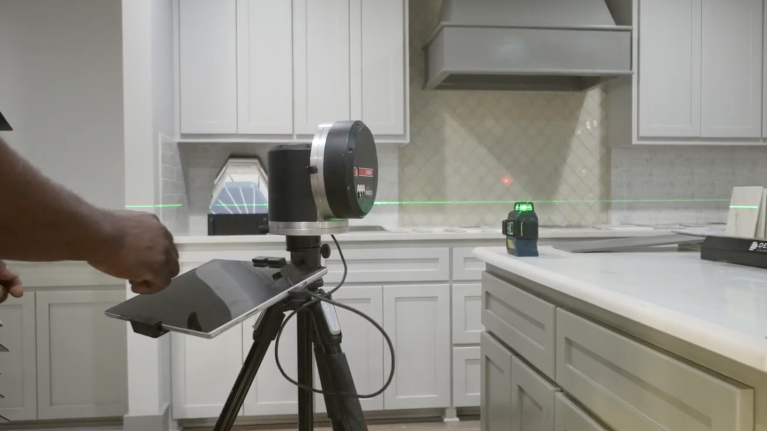 A person operating a laser scanner in a kitchen to measure and create a 3D map of the space, with kitchen cabinets, counters, and a laser beam visible.