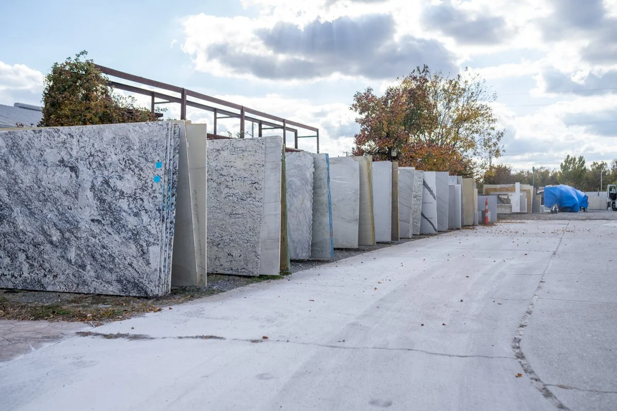 A row of large slabs of marble or granite stones standing upright outdoors on a cloudy day, with trees and a truck covered by a blue tarp in the background.