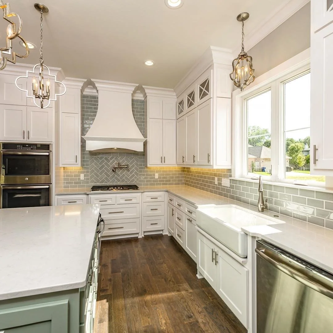 Bright kitchen with white cabinets, farmhouse sink, stainless steel dishwasher, double oven, gray backsplash, windows, and dark hardwood floors.