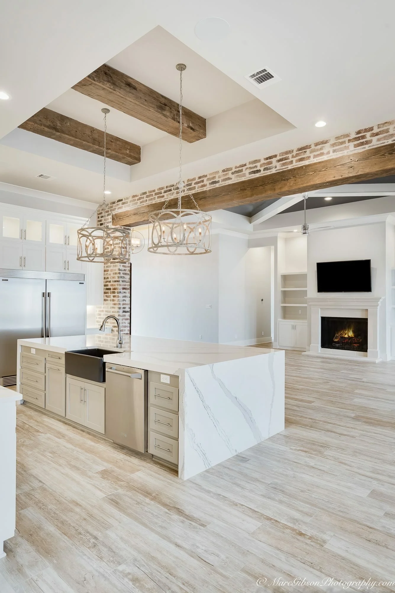 Open-concept kitchen and living area with white cabinets, marble island, brick and wood ceiling accents, and a fireplace with a mounted TV.