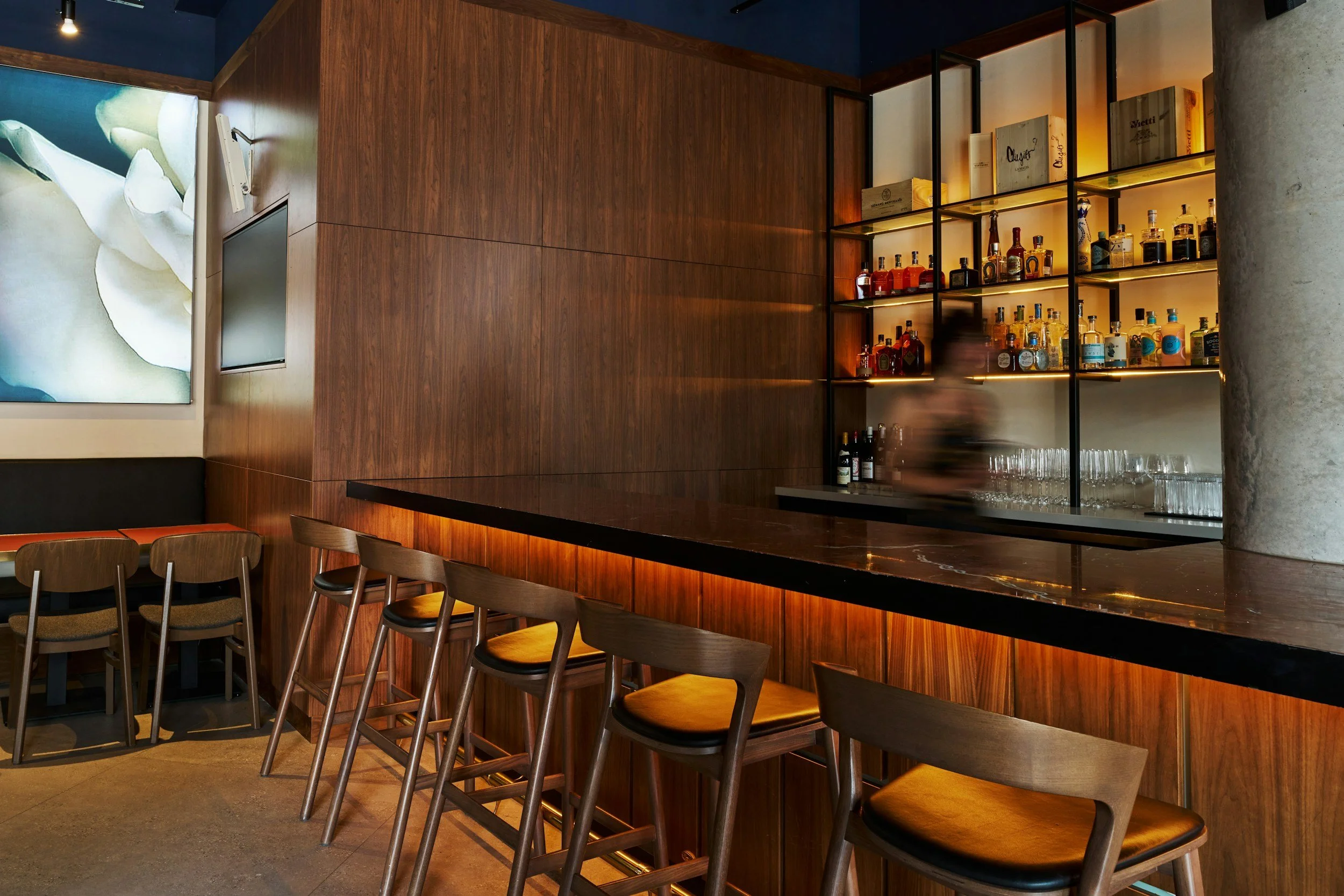 Empty bar with wooden counter and bar stools, backlit liquor shelves, blurred person behind the bar, modern interior decor.