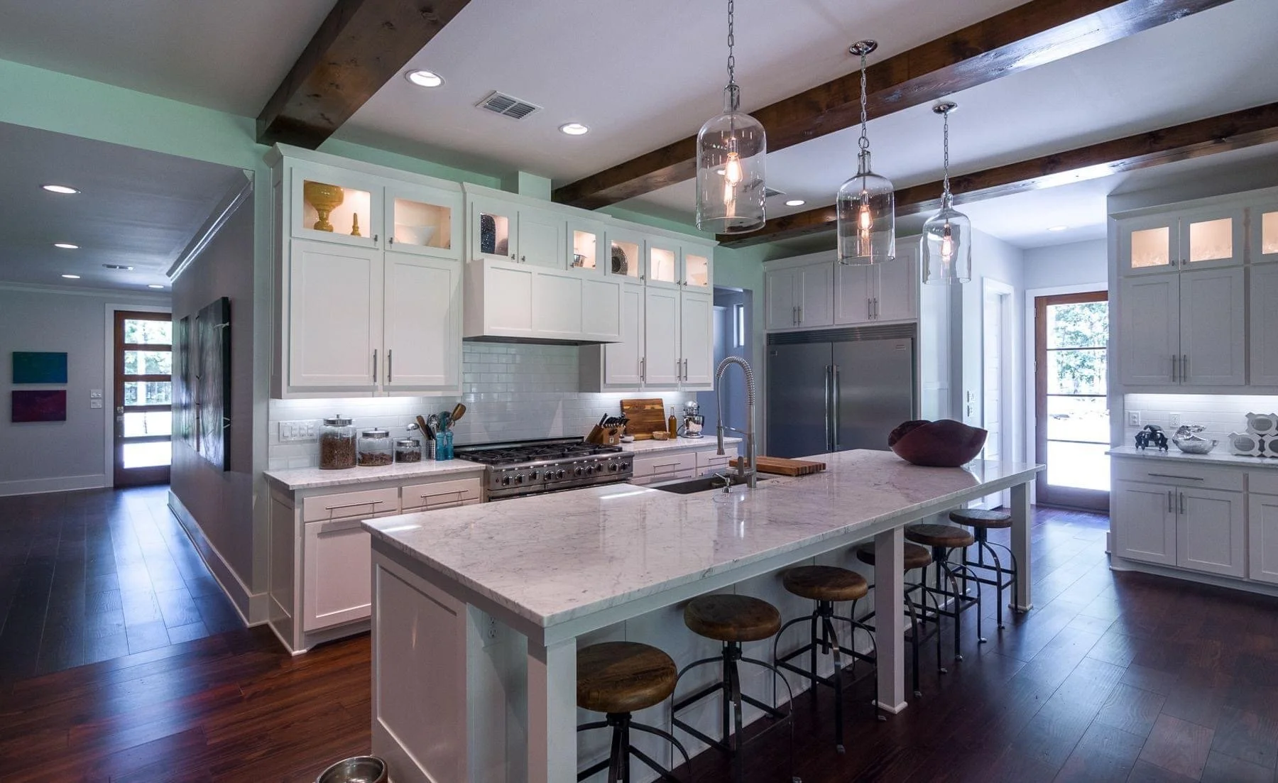 Modern kitchen with white cabinets, marble island, and wooden beams on the ceiling. Pendant lights hang over the island, and there is a window and door outside bringing in natural light.