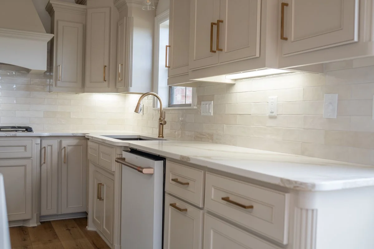 Modern kitchen with white cabinets, gold handles, marble countertop, and a beige brick backsplash. Includes a small sink with a gold faucet, under-cabinet lighting, and electrical outlets.