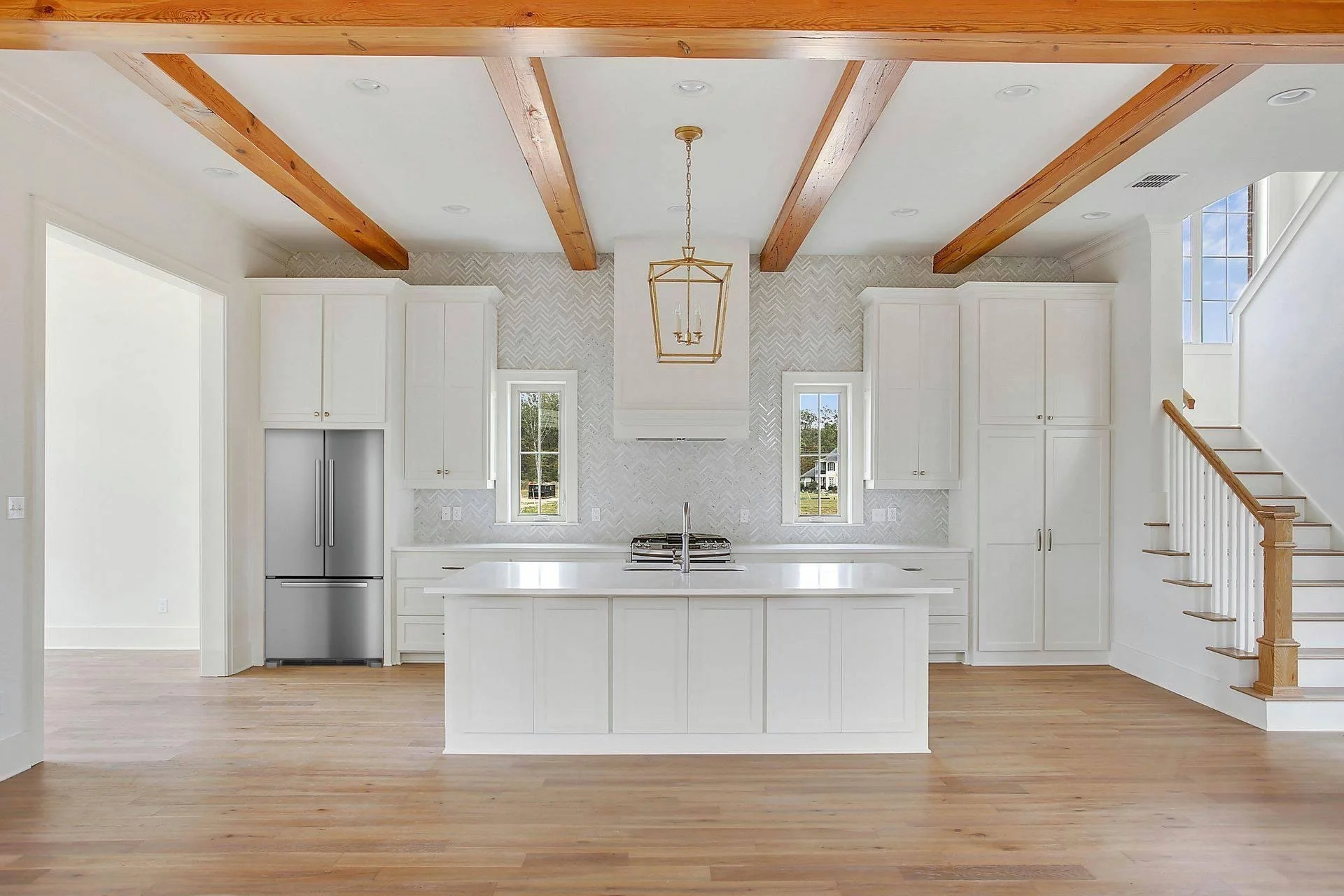 Modern kitchen with white cabinets, a central island, stainless steel refrigerator, two small windows, wooden ceiling beams, and a staircase on the right side.