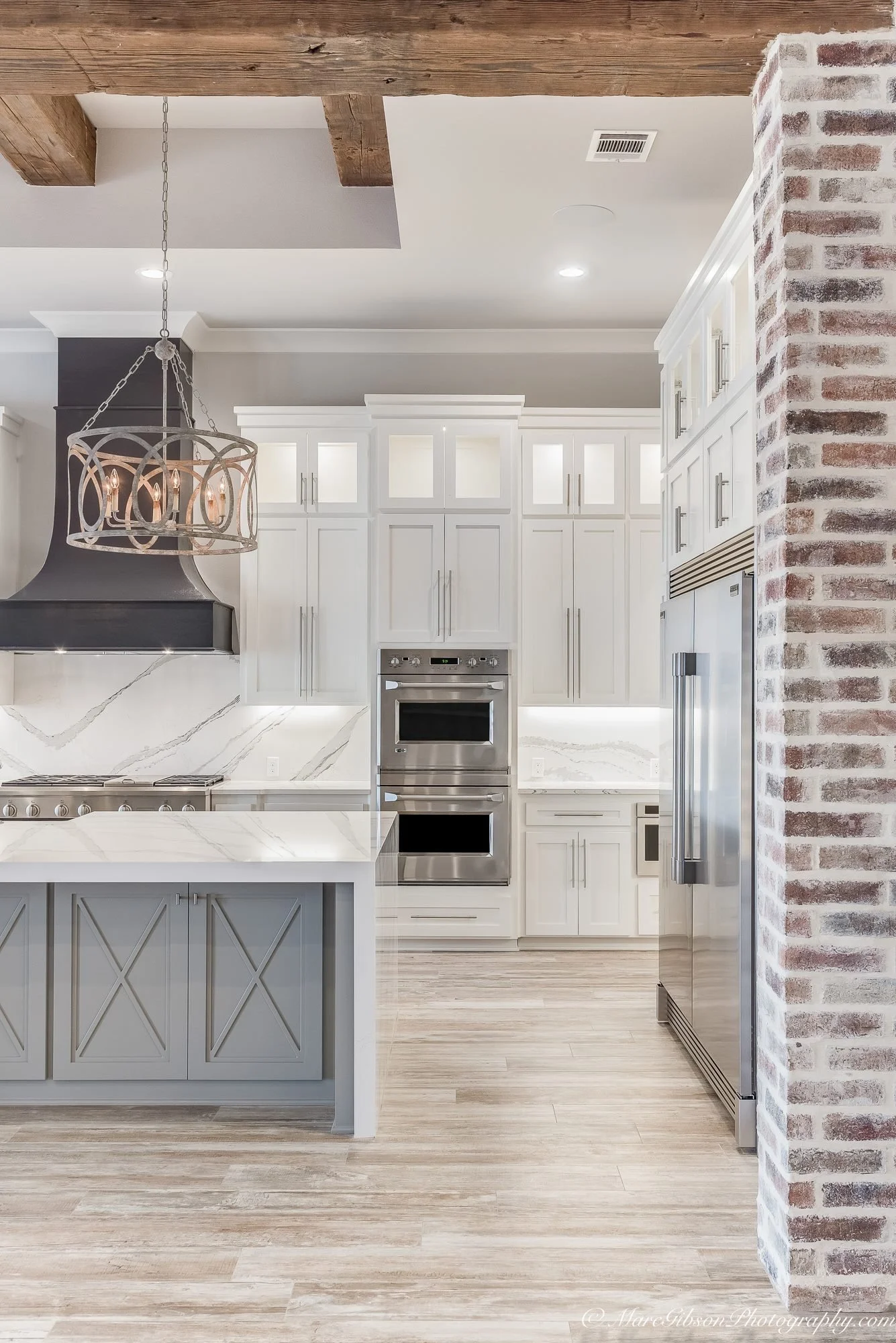 Modern kitchen with white cabinets, stainless steel appliances, a marble backsplash and island, exposed wooden beams, and an exposed brick wall.