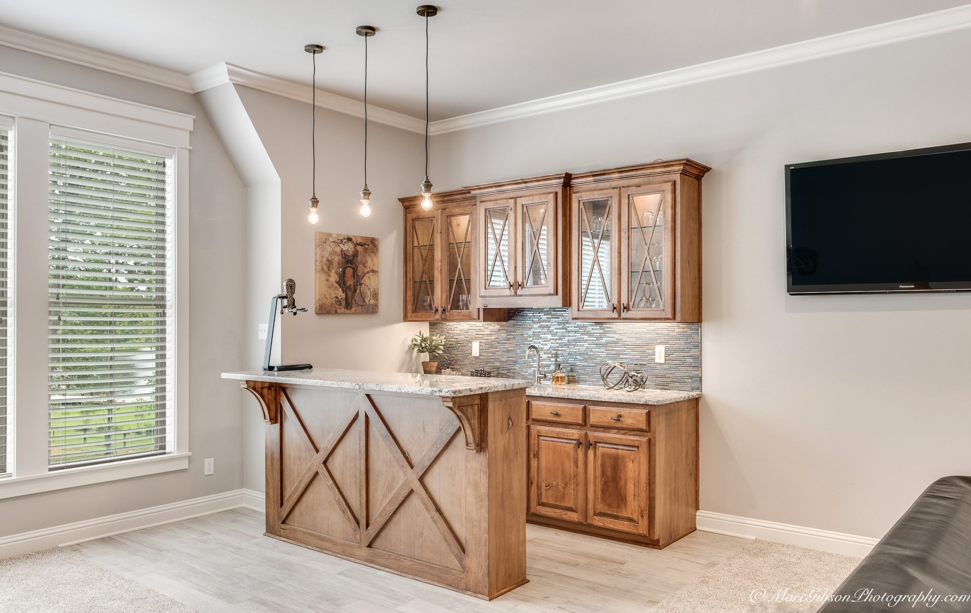 A kitchen with wooden cabinets, granite countertop, and a kitchen island, hanging pendant lights, a window with blinds, and a wall-mounted TV.