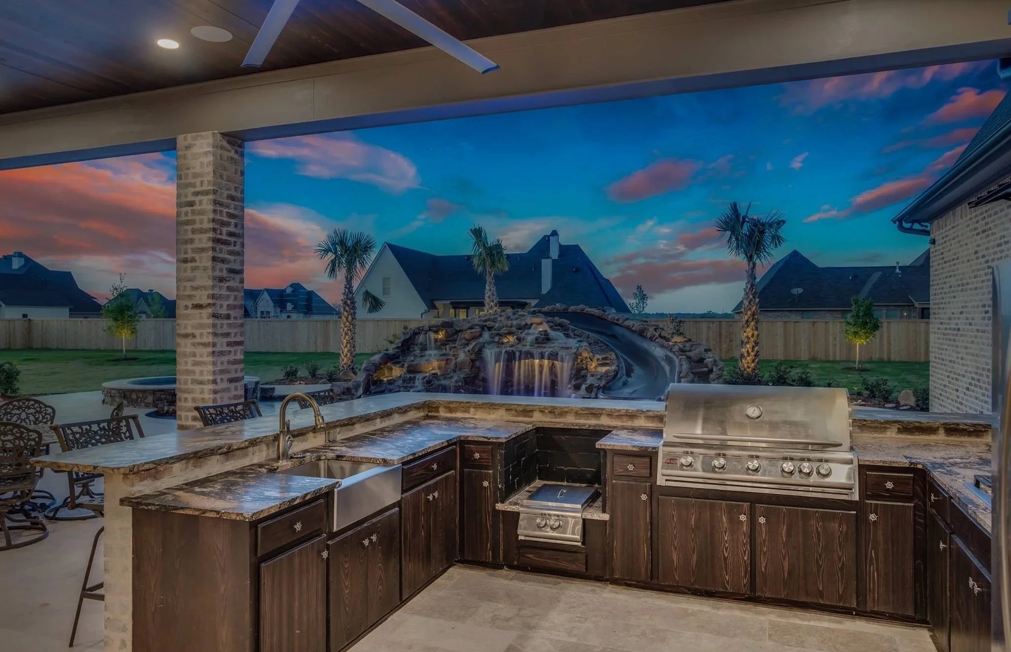 Outdoor kitchen with a grill, dark wood cabinets, granite countertops, and a scenic backyard view featuring a waterfall and palm trees during sunset.