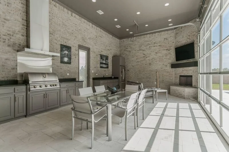 Modern indoor living space with a brick wall, large windows, a dining table with six chairs, a stainless steel barbecue grill, and a television mounted above a fireplace.