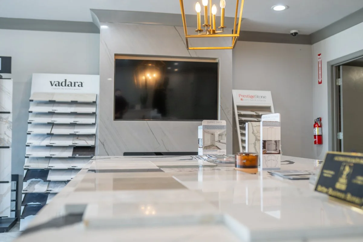 Interior of a showroom with display racks of tile samples, a large flat-screen TV on the wall, and a modern chandelier hanging from the ceiling.