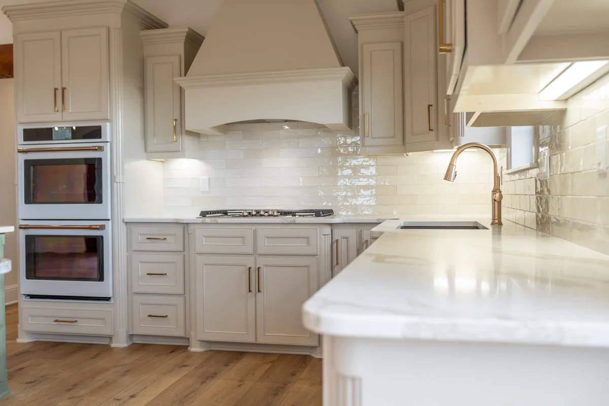 Modern kitchen with white cabinetry, a double wall oven, a gas stove, a white countertop, and a gold faucet over a black sink. Light hardwood floors and a white glossy tile backsplash complete the scene.