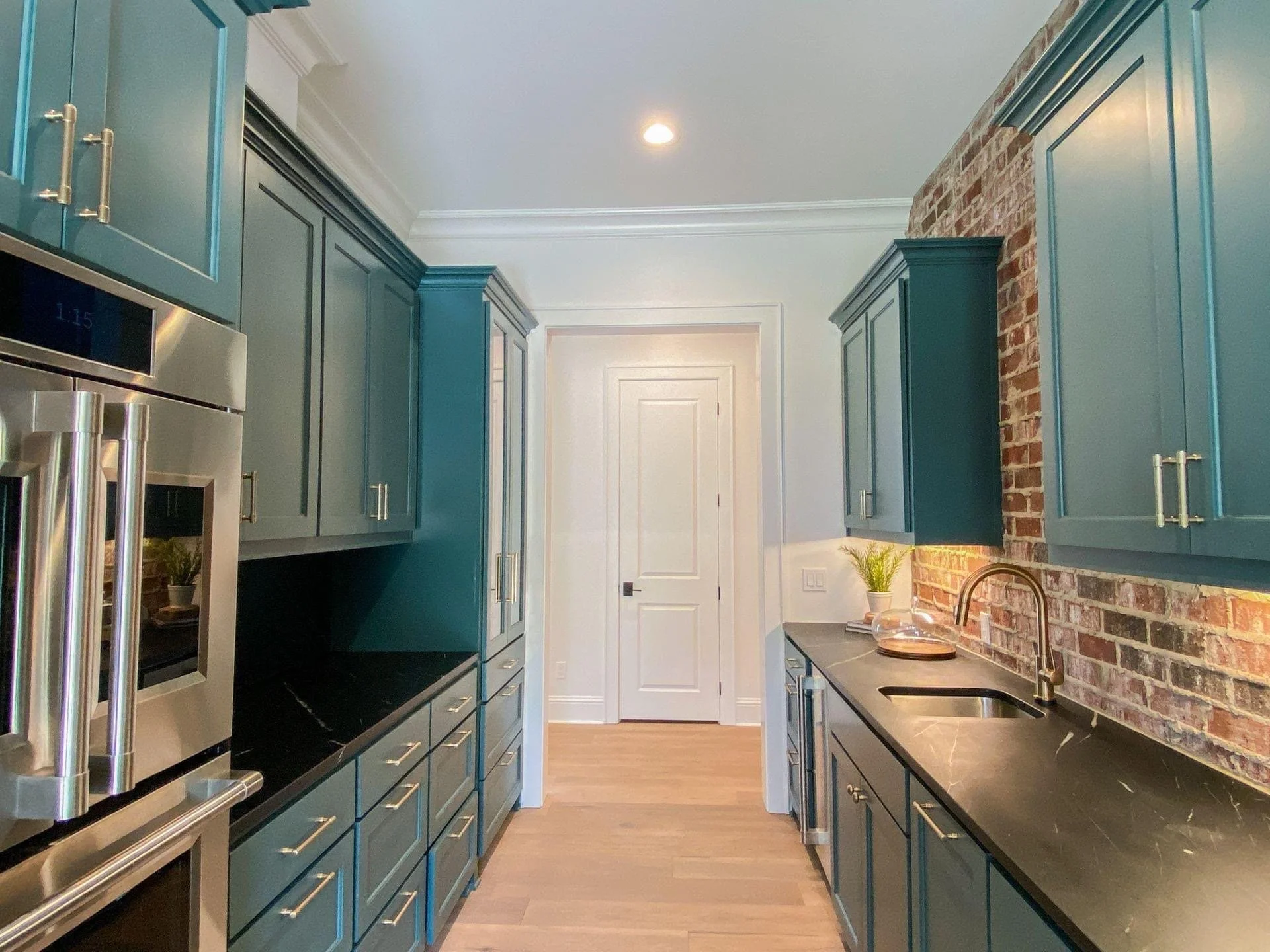 Modern kitchen with teal cabinets, black countertops, stainless steel oven, brick wall, wooden floor, and potted plant.