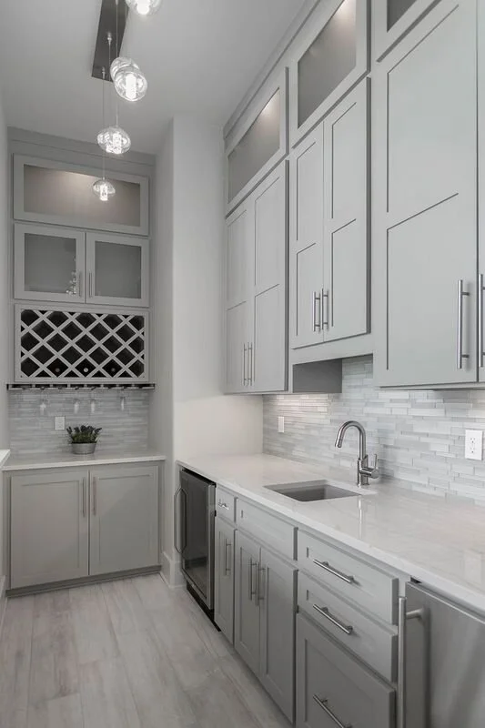 Modern kitchen with light gray cabinets, white marble countertops, and a gray backsplash. There are upper and lower cabinets with sleek handles, a small built-in mini fridge, and a single-basin sink with a modern faucet. A potted plant is on the coun