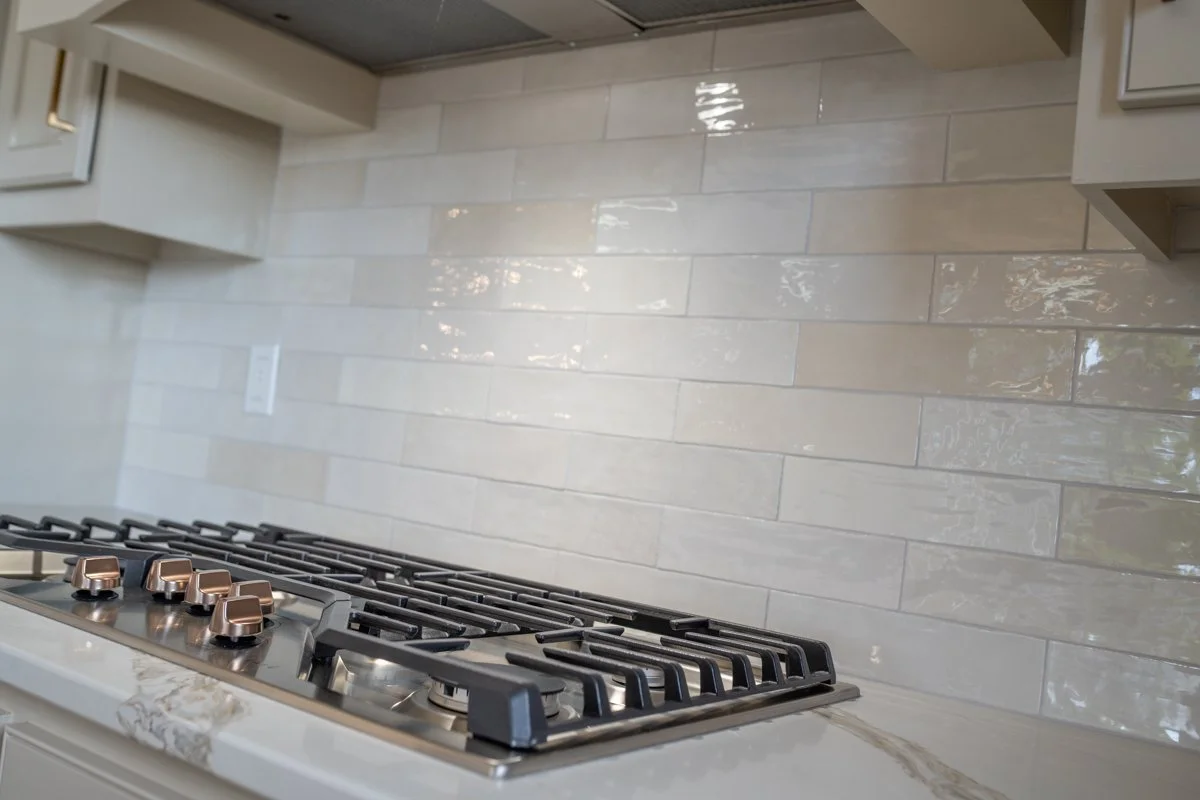 Kitchen stovetop with black grates and four copper-colored control knobs, beige marble countertop, and light-colored tile backsplash.