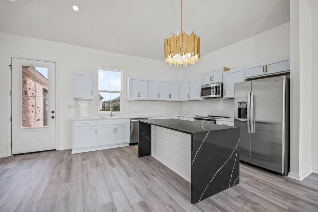 Modern, white kitchen with light wood flooring, stainless steel appliances, and a black marble island with white veining, hanging pendant light, and a door leading outside.