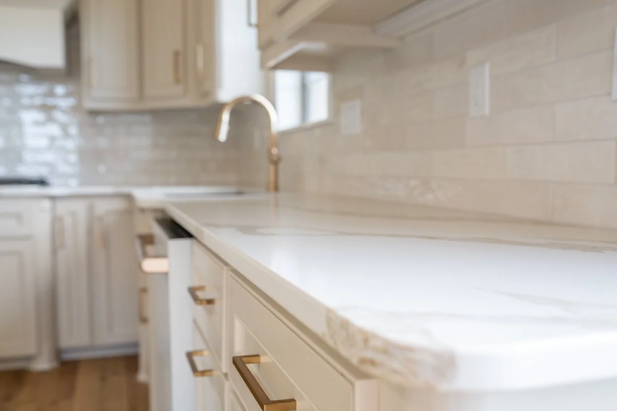 A modern kitchen with light-colored cabinets, a white countertop, a gold faucet, and a beige tile backsplash.