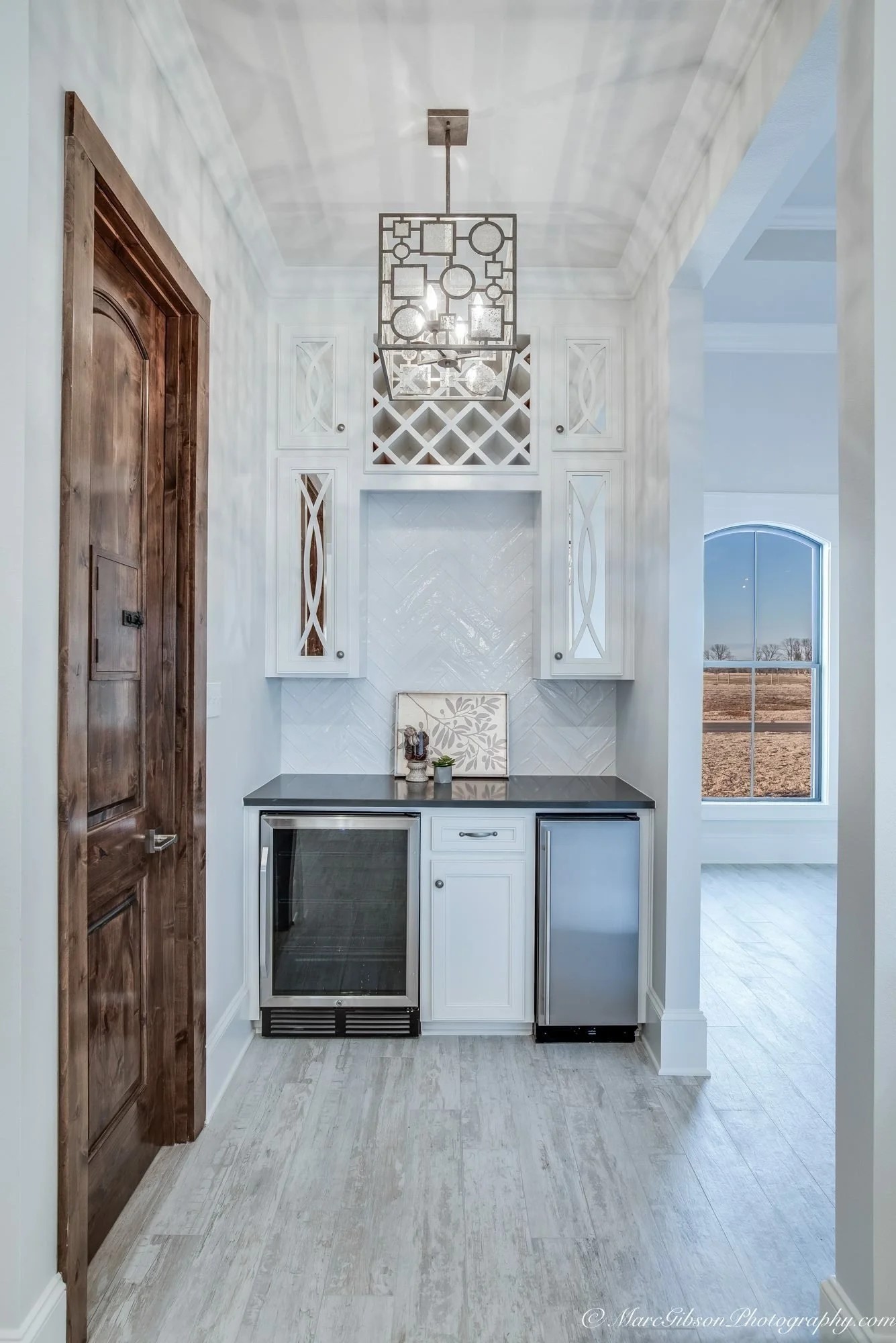 A small built-in wet bar area with white cabinetry, a black countertop, and a wine fridge. Decor includes small plants and a framed leaf print. There is a window with a view of a field outside. An elegant, modern chandelier hangs from the ceiling.