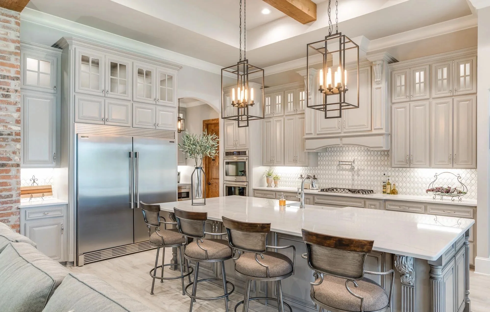 A modern kitchen with beige cabinetry, a large white island with barstools, stainless steel refrigerator, double wall oven, pendant lighting, and a brick accent wall.