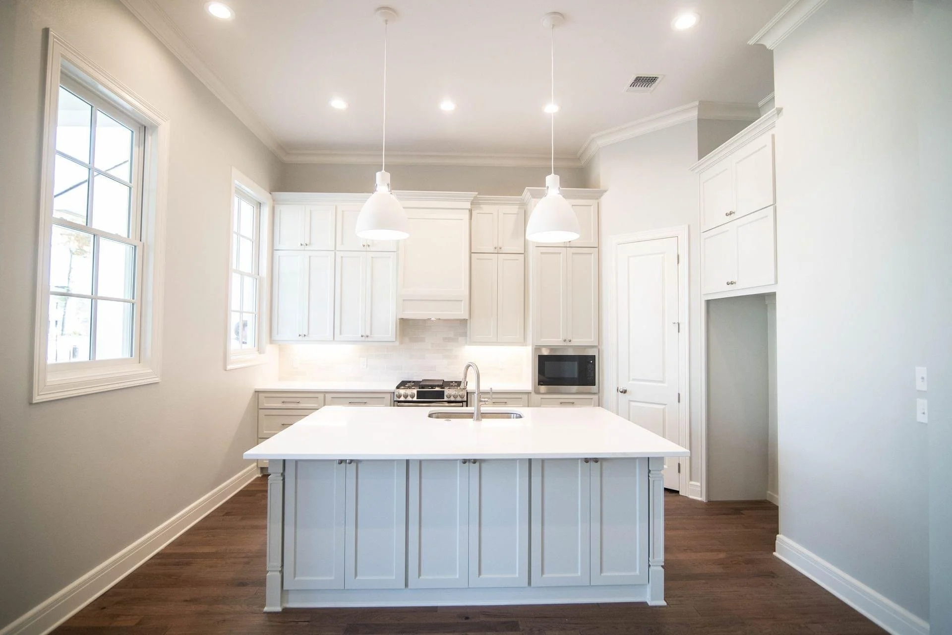 Bright kitchen with white cabinets, a large island with a sink, and two pendant lights. Has two windows, a stove, microwave, and hardwood floors.