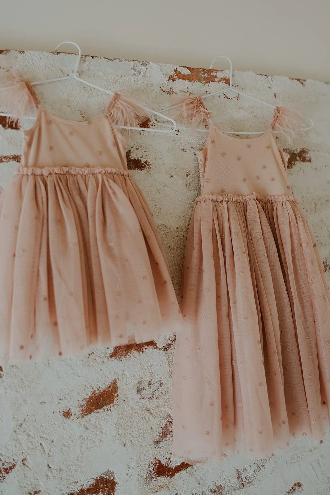 Two pink dresses with puffed sleeves and pleated skirts hanging on white hangers against a rough white brick wall.
