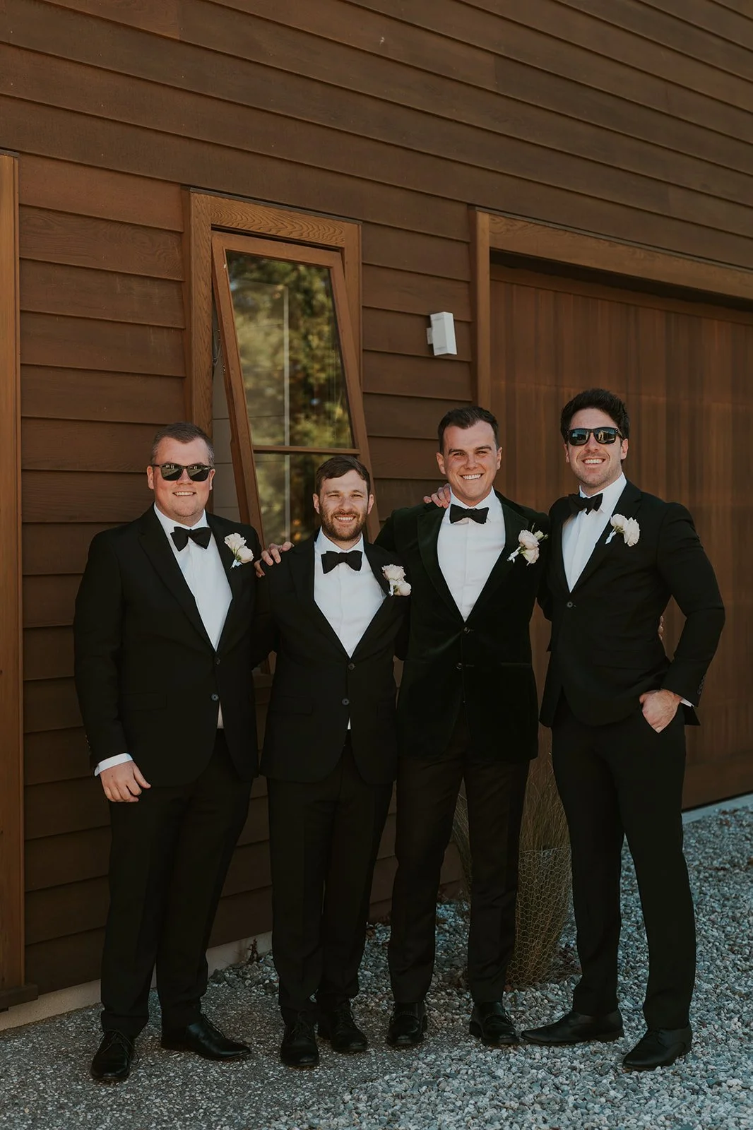 Four men in black tuxedos with white shirts and black bow ties, standing outside against a brown wooden building. They have boutonnieres on their lapels, appear to be at a wedding or formal event. Two are wearing sunglasses, smiling, with arms around each other.