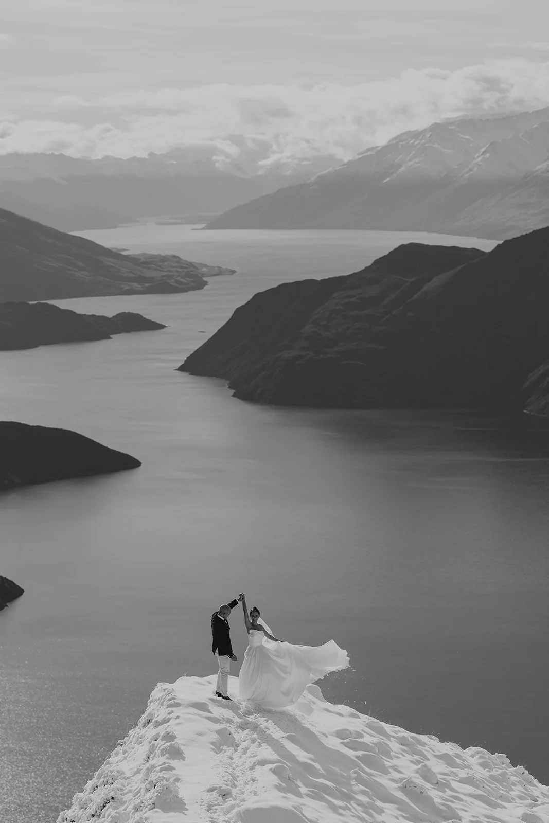 A bride and groom dancing on a snowy hilltop overlooking a mountain lake in black and white.