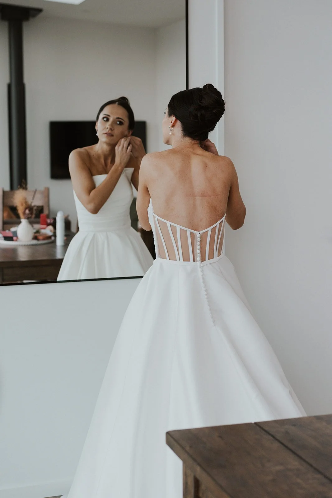 Bride with dark hair in an updo wearing a white wedding dress with a lace back, looking at herself in a mirror while adjusting earrings.