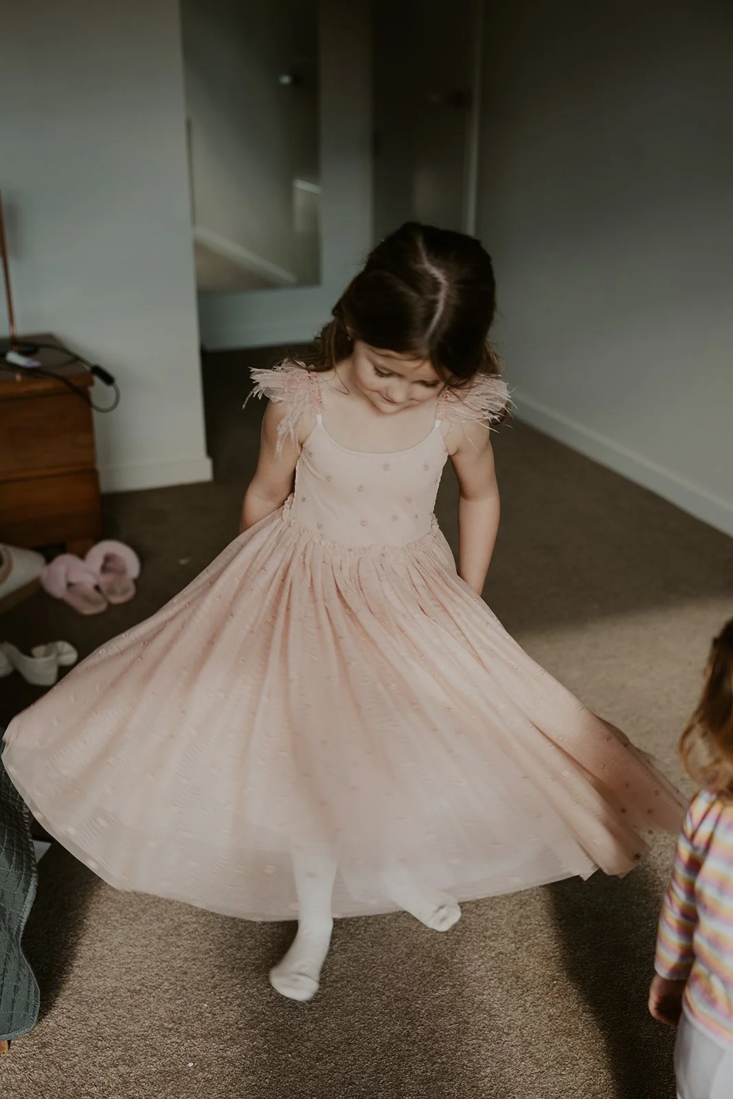 A young girl wearing a pink dress with possibly tulle detailing at the shoulders, looking down at her dress, as she twirls or spins, causing her dress to flare out around her. She is standing on a carpeted floor in a room.