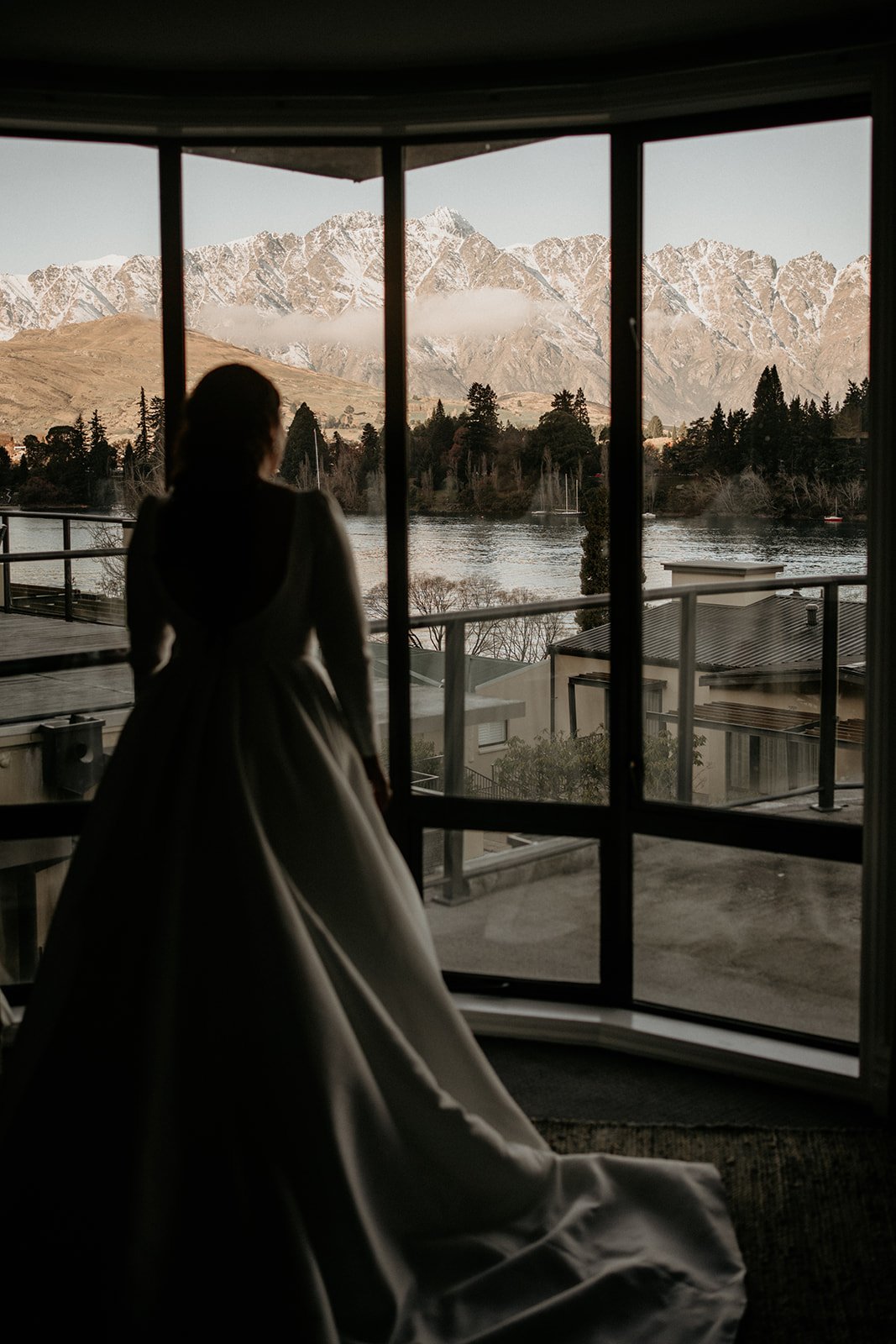 A woman in a long dress standing indoors by a large window, gazing at a mountain landscape with snow-capped peaks, a lake, and trees.