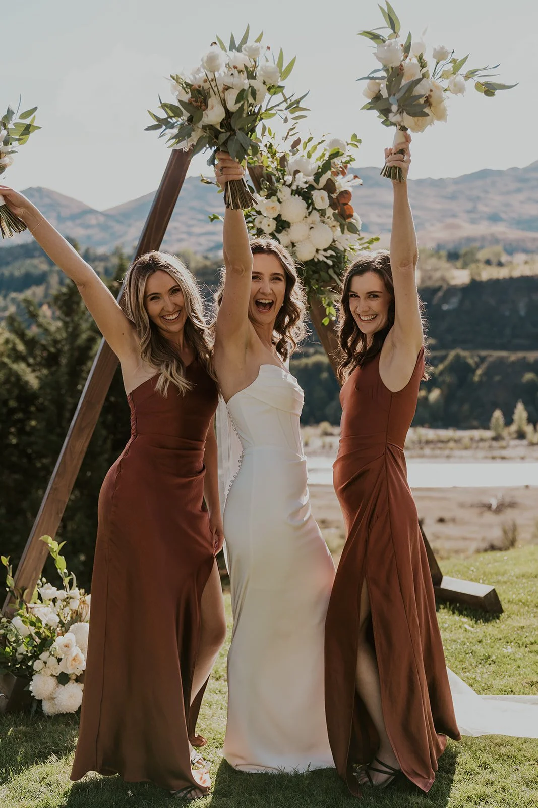 Three women at a wedding, one in a white wedding dress and two in brown bridesmaid dresses, celebrating outdoors with arms raised and holding bouquets, smiling happily, with a scenic mountain backdrop and floral arch.