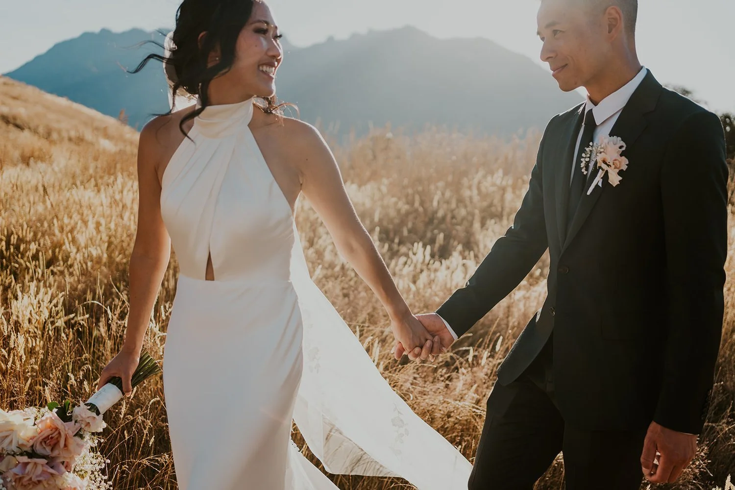 A bride and groom holding hands in a field with mountains in the background during sunset.