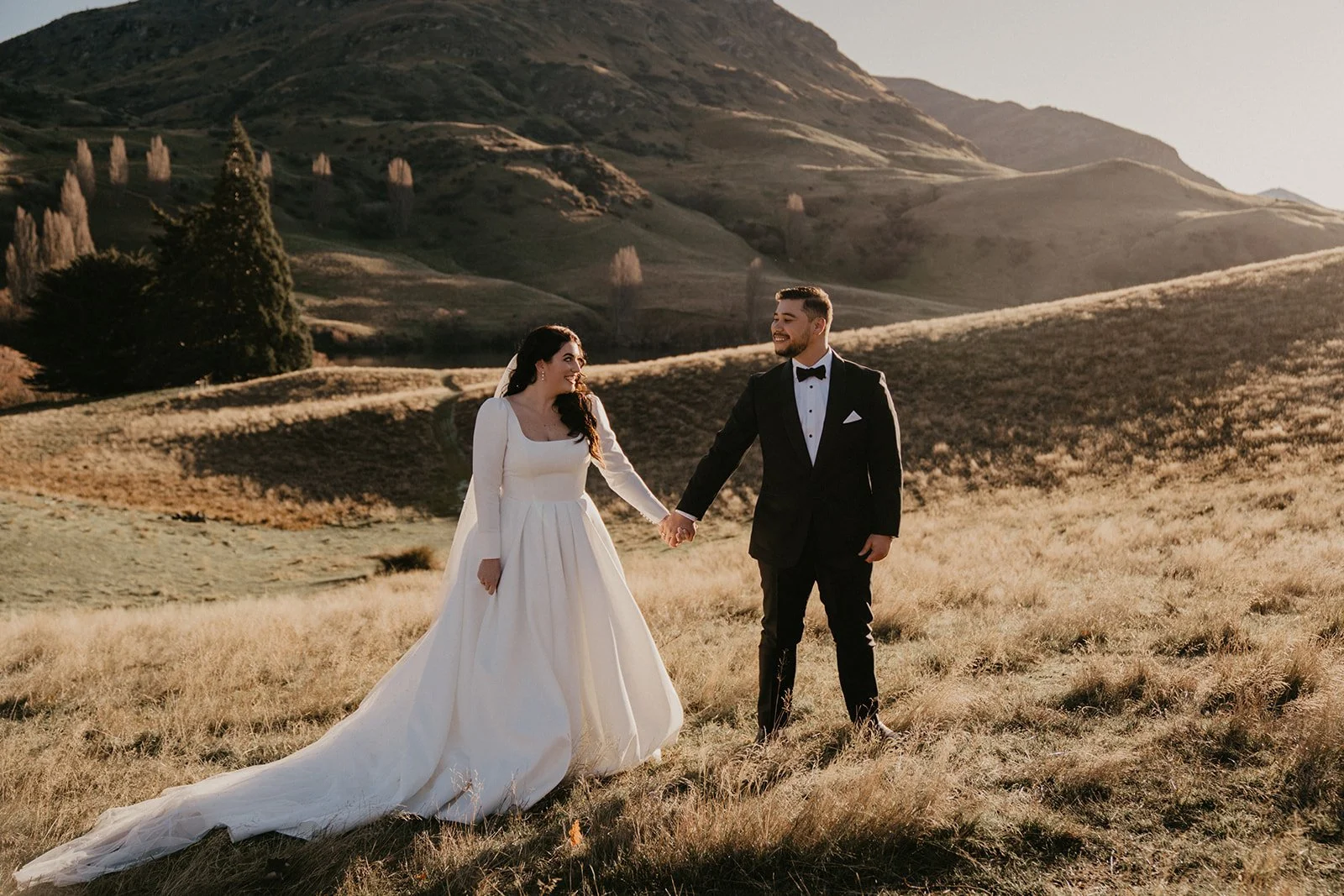 A bride and groom holding hands and smiling at each other in a grassy hillside landscape during sunset, with rolling hills and sparse trees behind them.