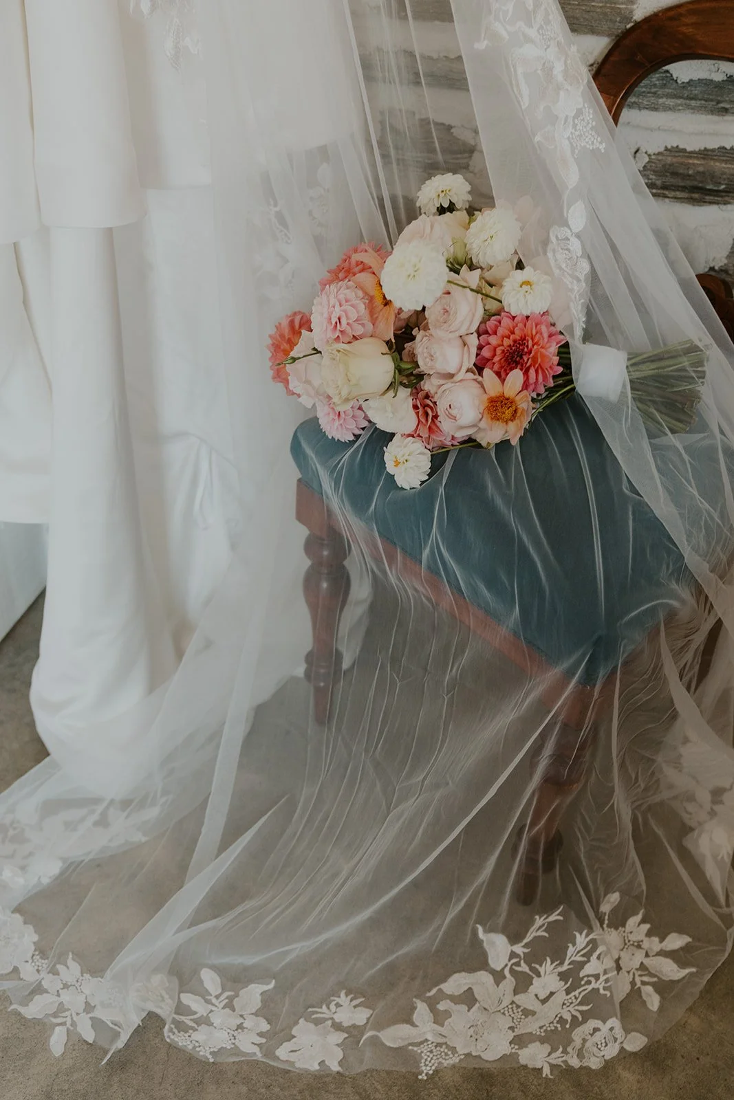 A bouquet of pink, peach, and white flowers resting on a small wooden table, covered with a sheer, embroidered fabric with floral patterns, set against a rustic wall.