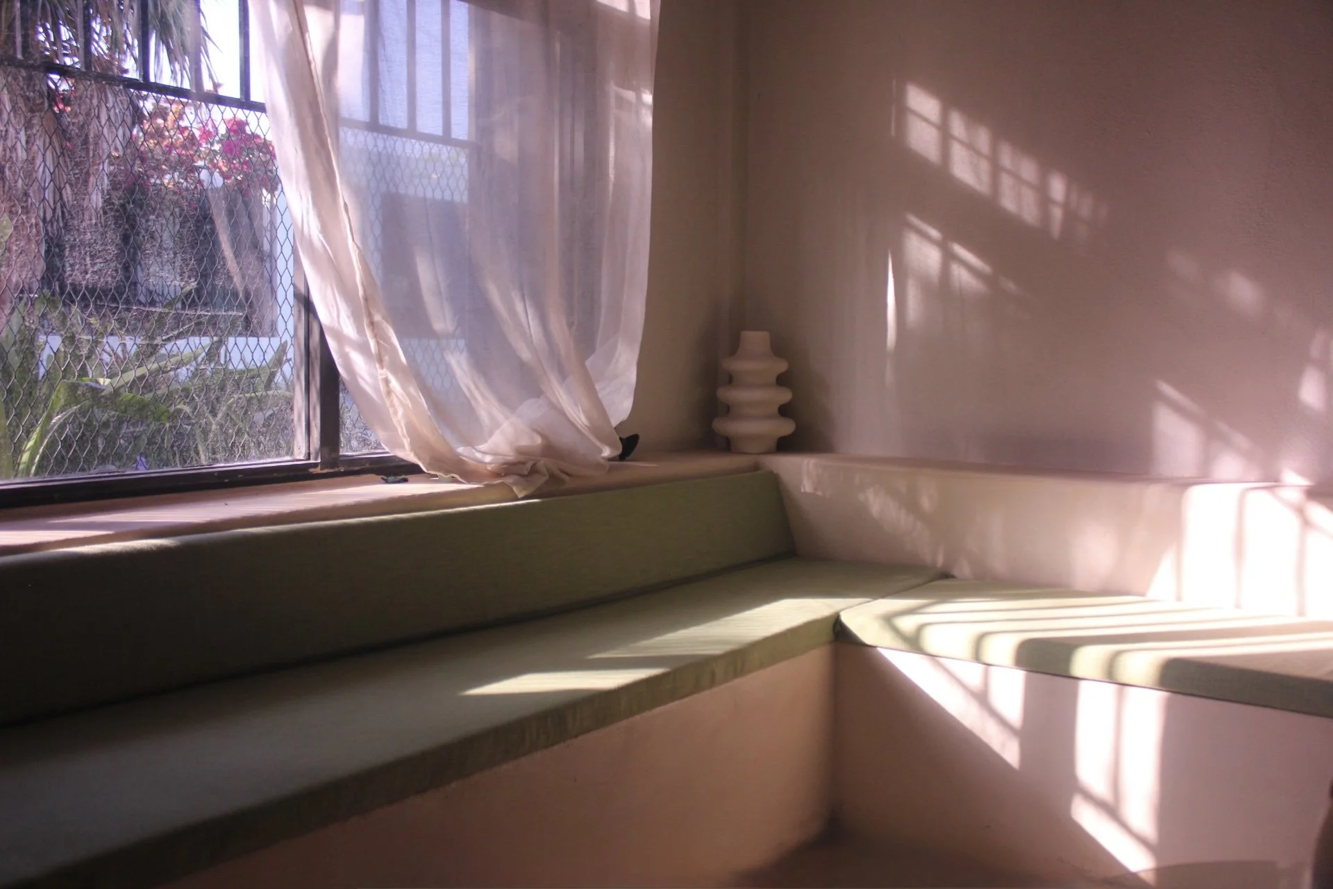 Sunlight streams through a window with sheer curtains, casting shadows on the beige wall. There is a green cushioned bench along the window and a small, abstract white table or sculpture in the corner.