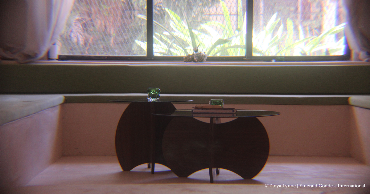 A small black side table with a rounded design, placed in front of a window with curtains. The table has two glass candles and some stacked books on it. Outside the window, a garden with green plants is visible.
