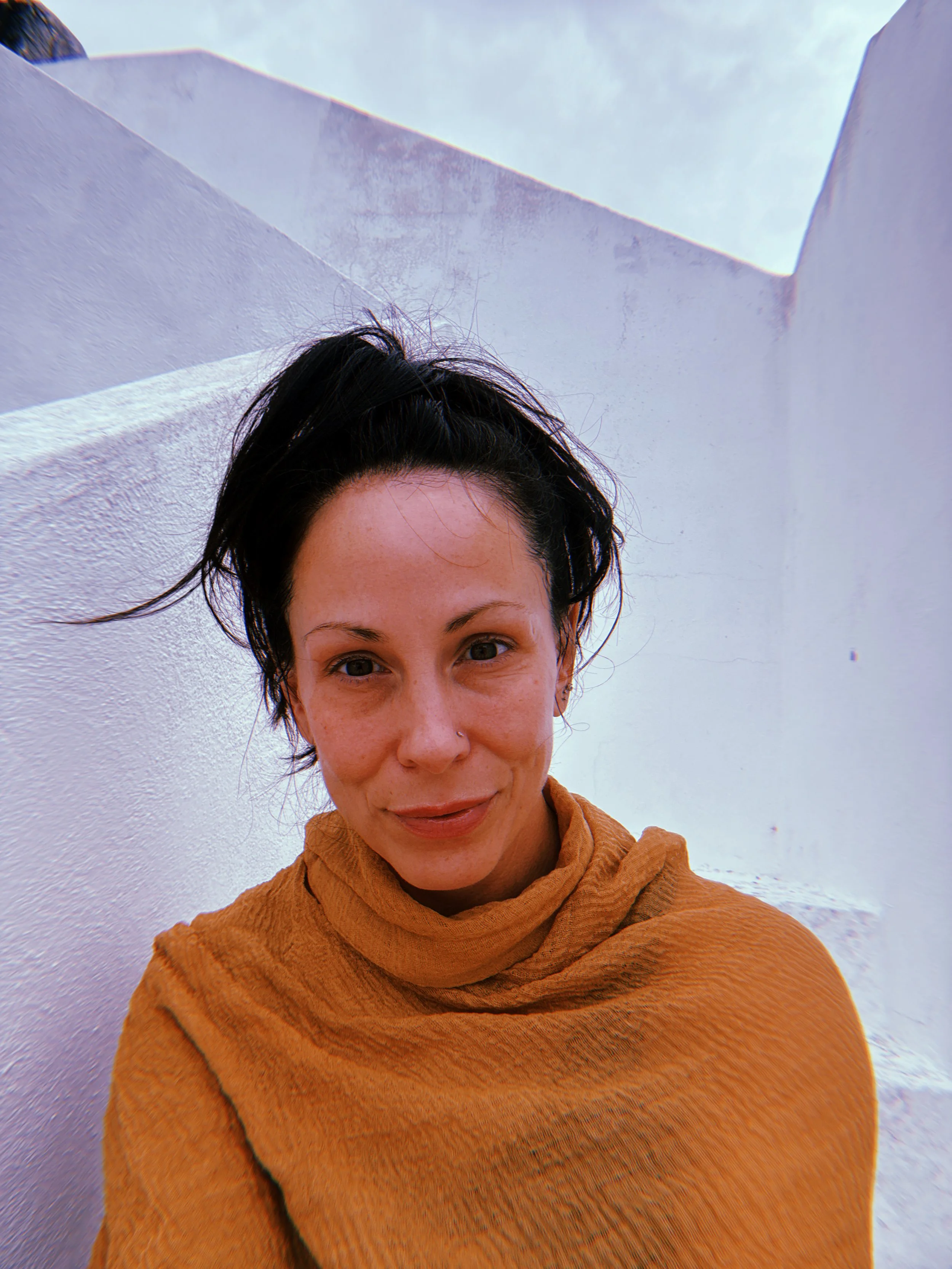 A woman with brown hair tied in a bun, wearing a brown scarf, standing against white walls with blue sky visible above.