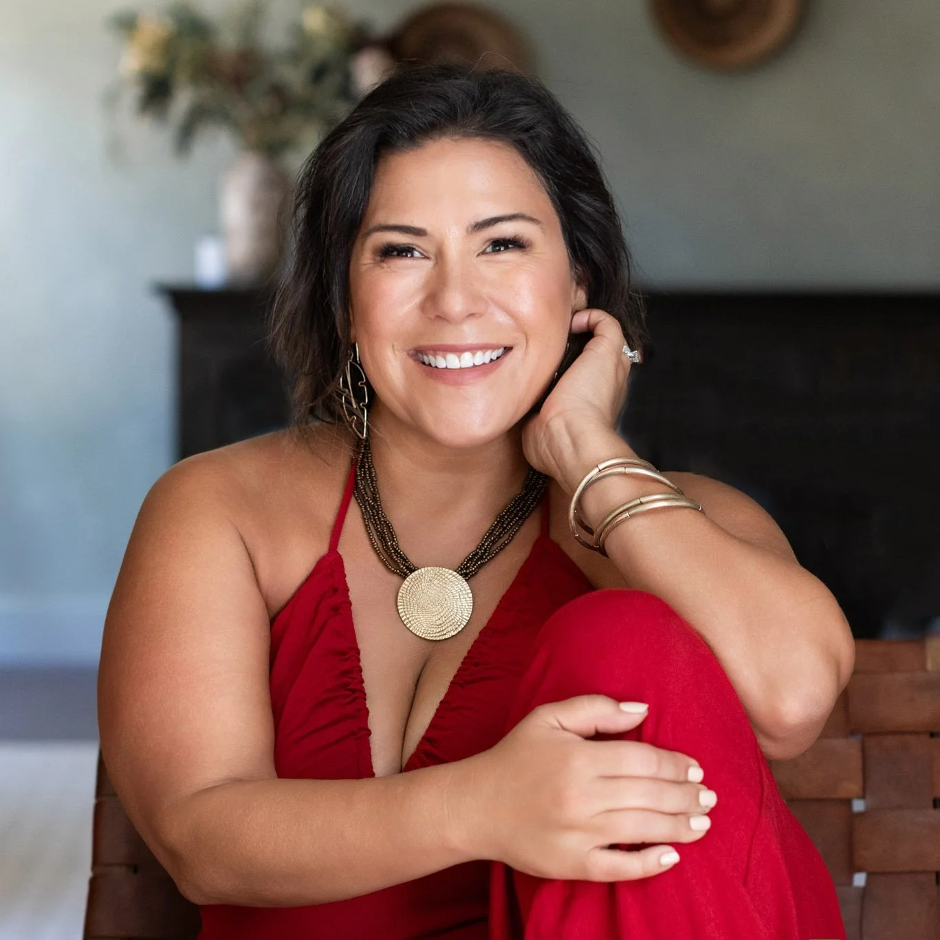 A smiling woman with dark hair, wearing a red dress and jewelry, sitting on a bench in a cozy indoor setting.