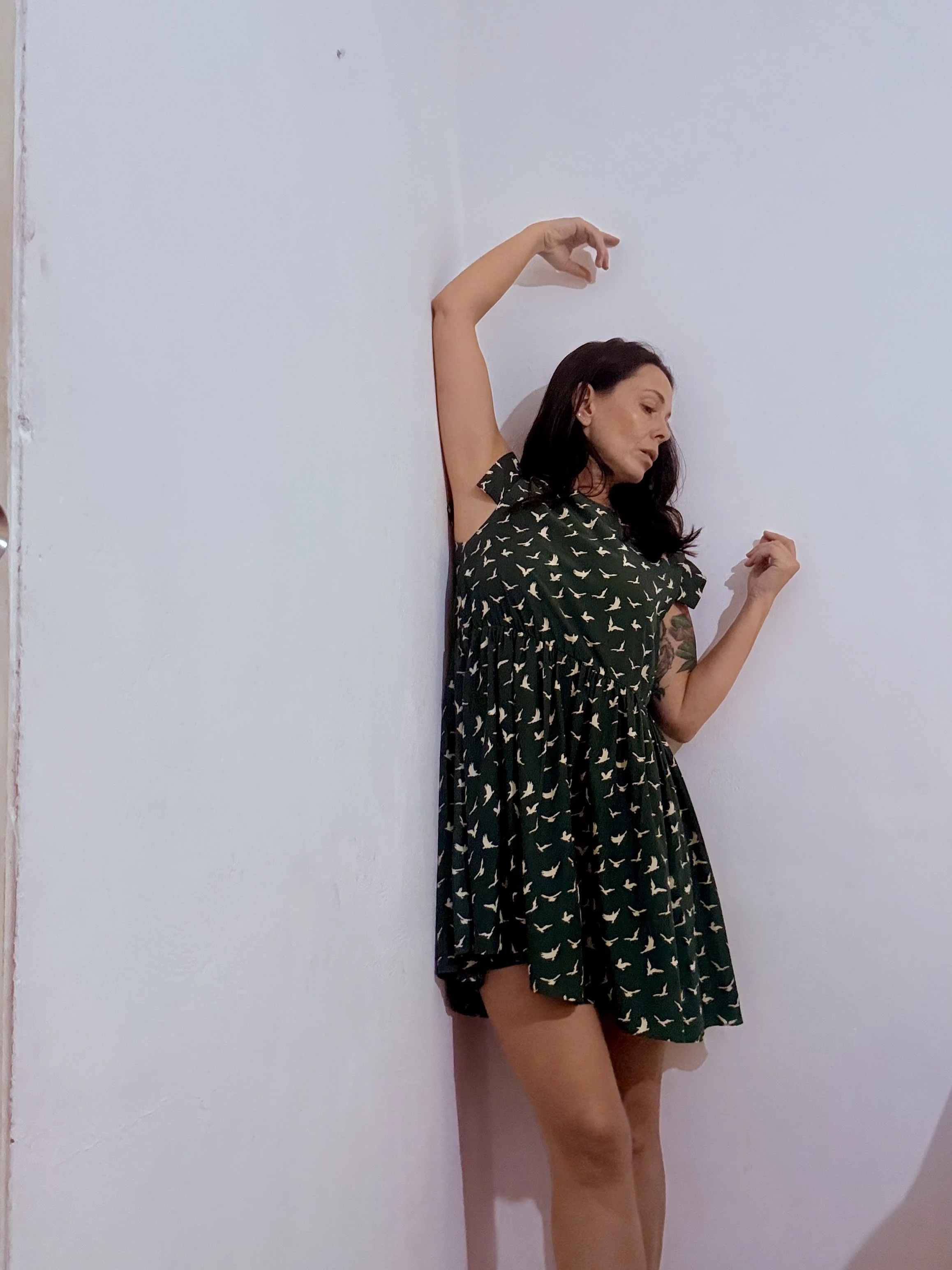 Woman with dark hair in a dress with a bird pattern, standing against a plain white wall, with her eyes closed and one arm raised above her head.