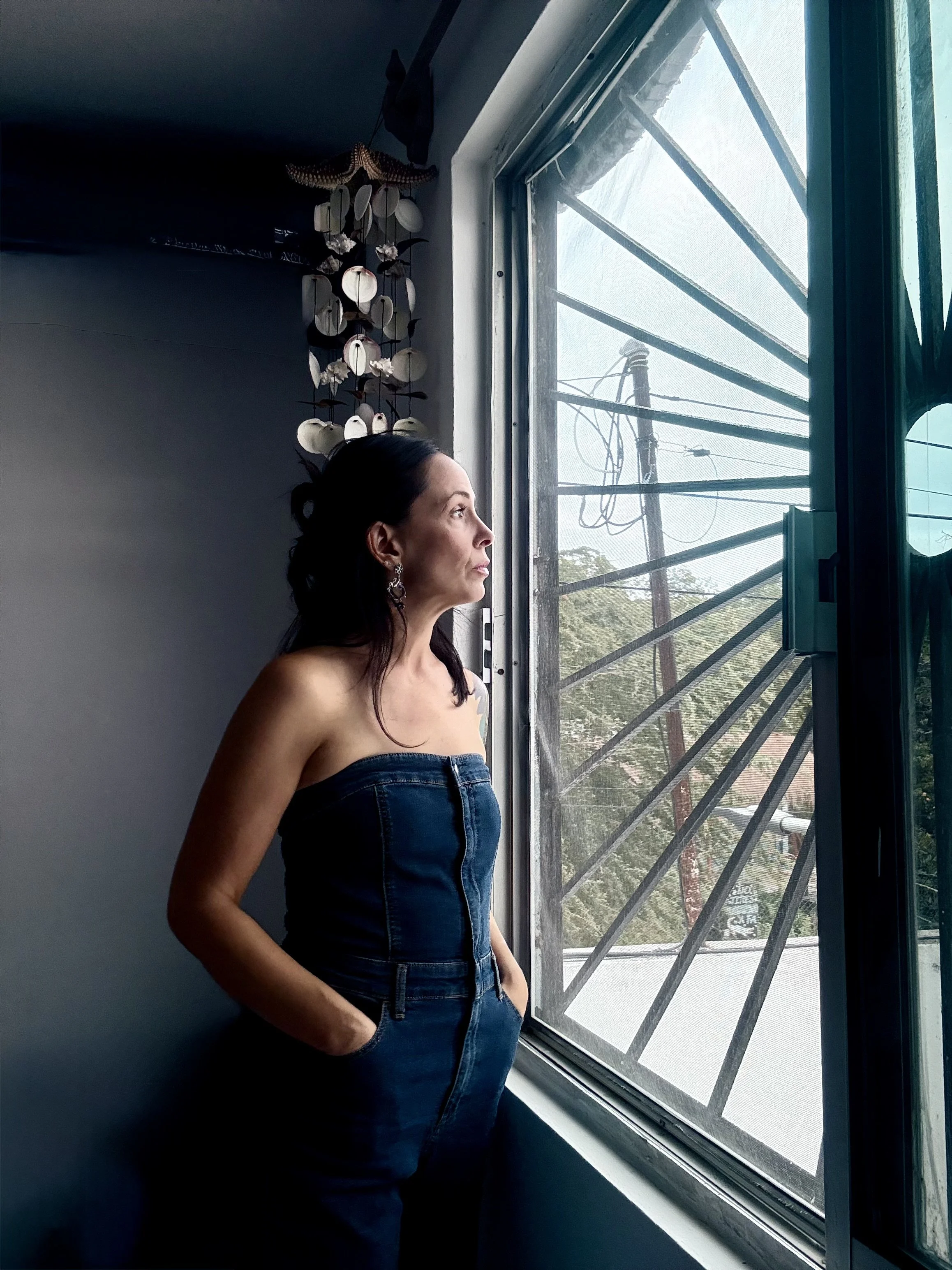 A woman with long dark hair wearing a strapless denim top and earrings stands by a window, looking outside with a contemplative expression.
