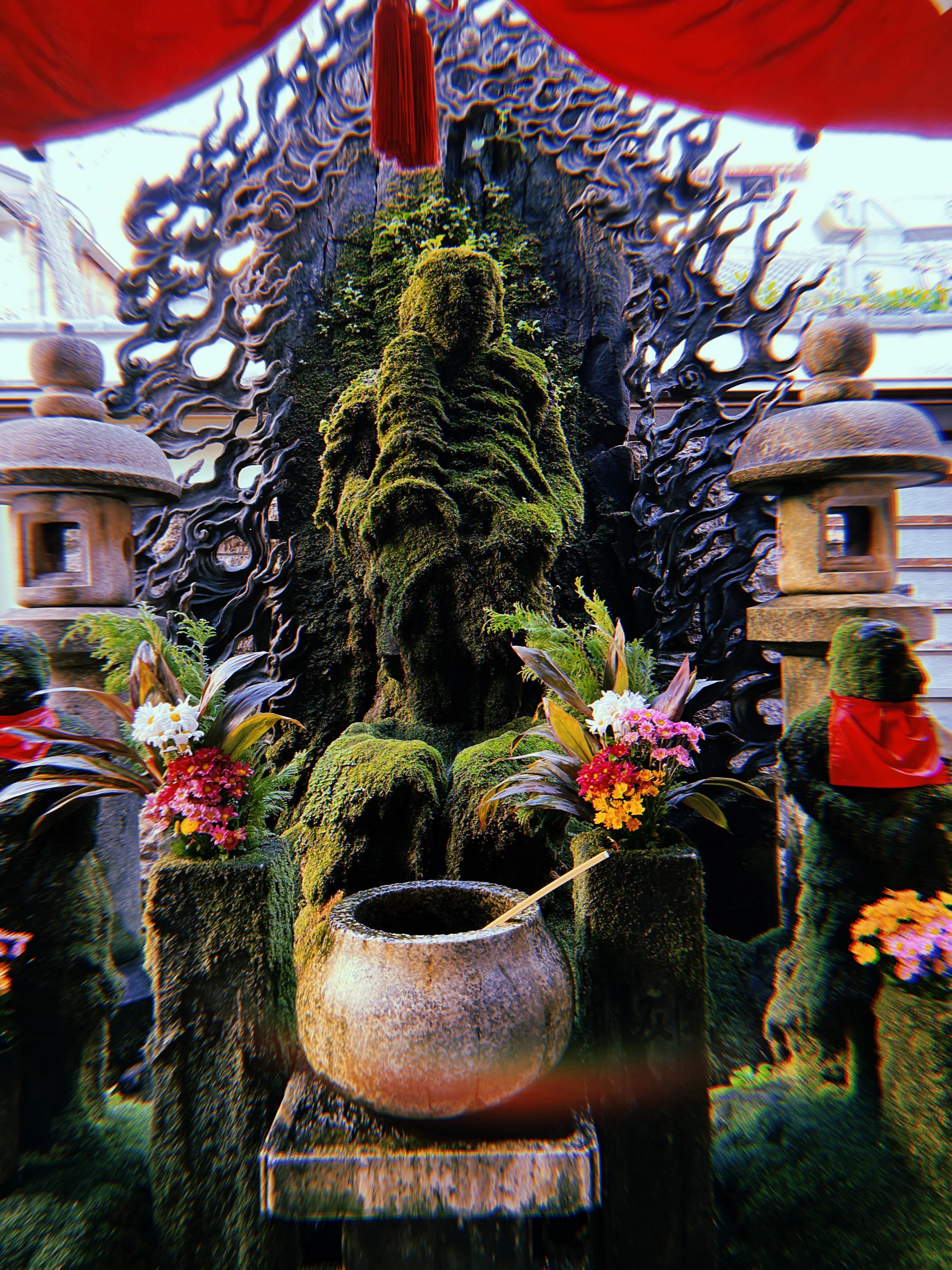 An ornate indoor altar with a moss-covered Buddha statue, floral arrangements, stone lanterns, and a large water basin, likely in a temple setting.