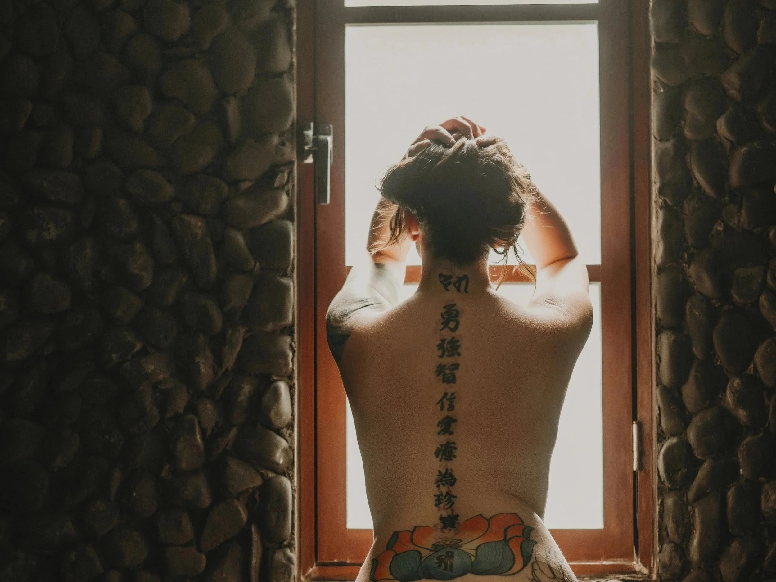 A person with tattoos on their back standing in front of a window, with their arms raised and hands on their head, framed by stone walls.