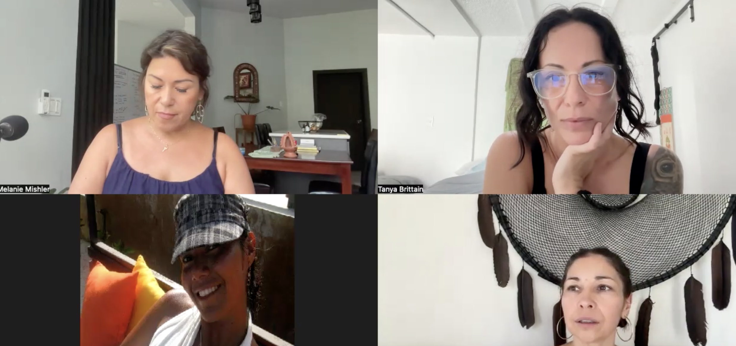 Four women participating in a video conference call. Top left: a woman with short brown hair wearing a navy sleeveless top, in a room with a white wall and a dining area. Top right: a woman with shoulder-length dark hair and glasses, resting her chin on her hand in a bright room with a bed. Bottom left: a woman wearing a plaid cap and earrings, outdoors with a background of fence and greenery, smiling and carrying a pole with a pillow. Bottom right: a woman with dark hair tied back, in front of a black hanging decoration with feathers, outdoors or in a light-colored indoor space.