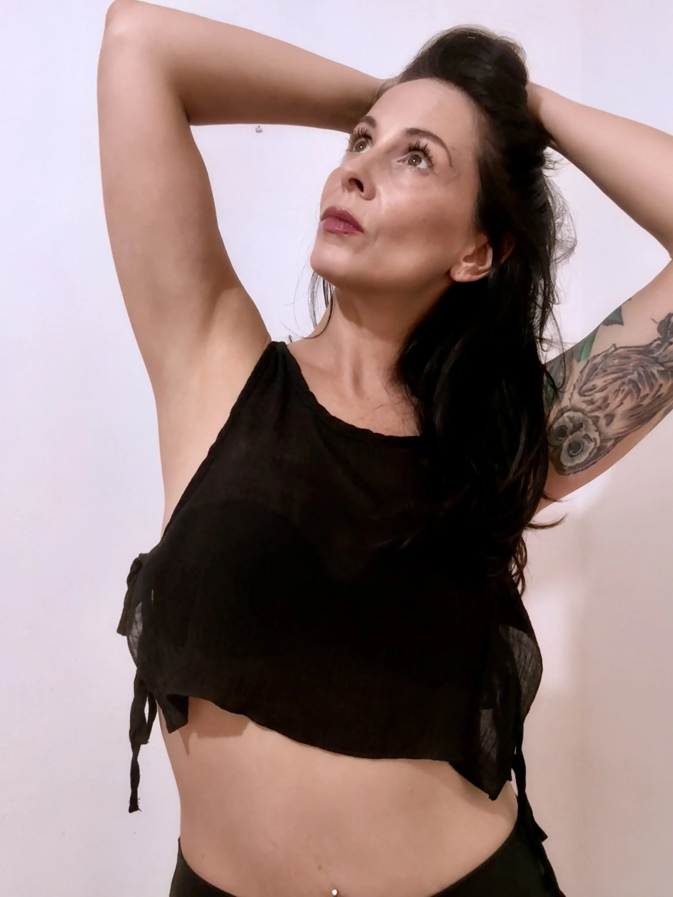 A woman with long dark hair, wearing a black sleeveless top, posing with arms raised and hands behind her head, looking upwards, with a tattooed arm visible, standing against a plain white background.
