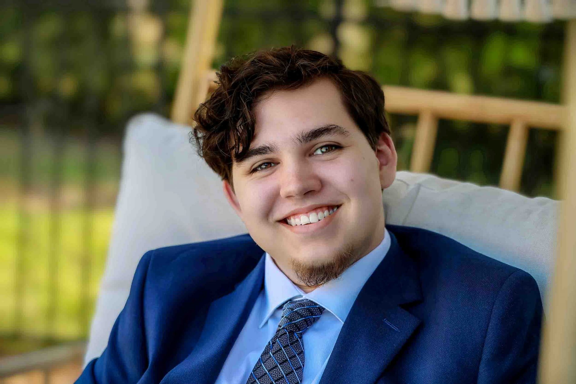 Senior Prom Dinner Event A young man with dark curly hair, light blue eyes, and a goatee smiling while sitting outdoors at Pace Prom Dinner Event. He is wearing a dark blue suit, blue dress shirt, and tie. 