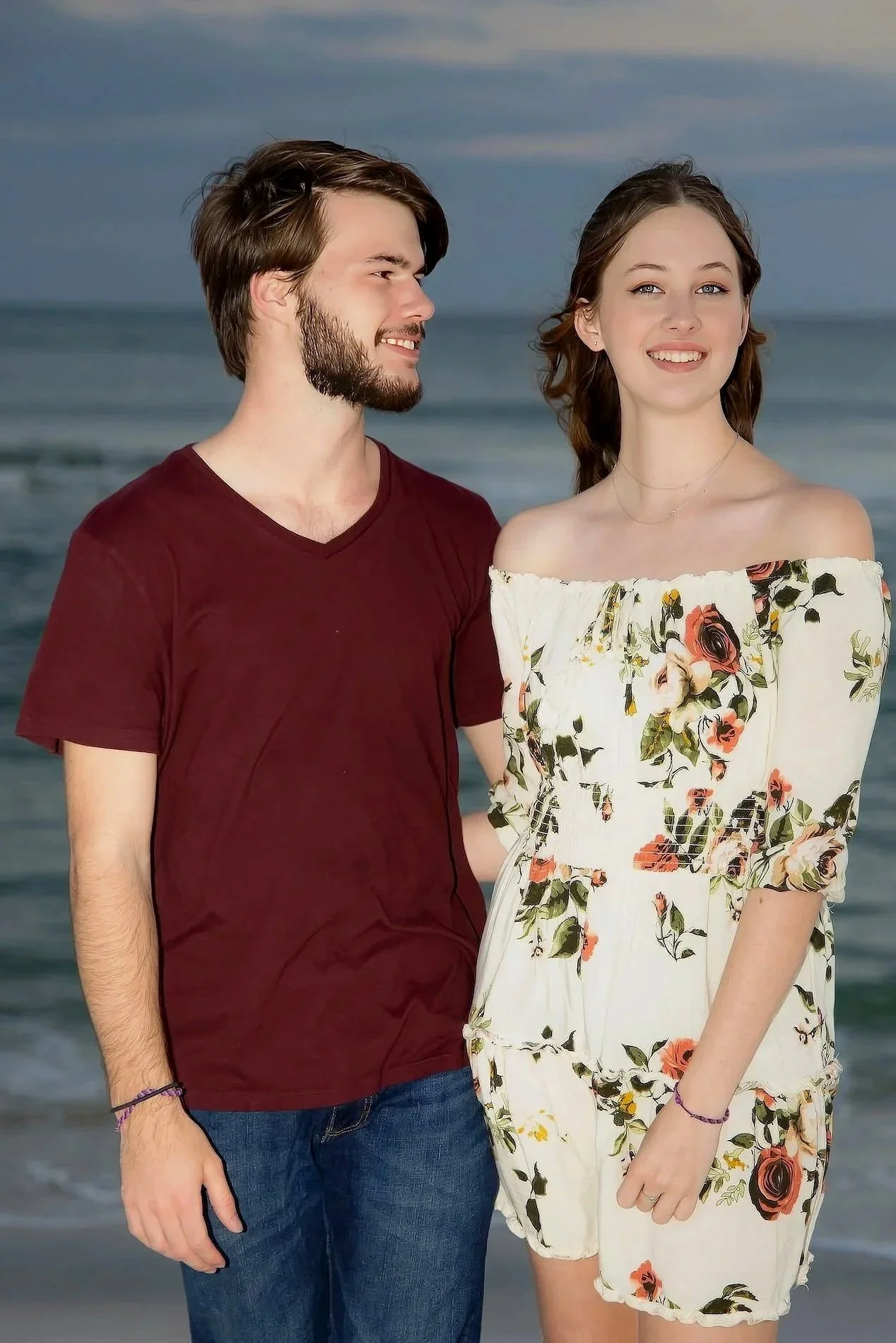 Blue hour Pensacola Beach Portrait A young couple standing on the beach, with the ocean and sky in the background. The man has brown hair and a beard, wearing a maroon T-shirt and jeans. The woman has long brown hair and is wearing an off-shoulder fl
